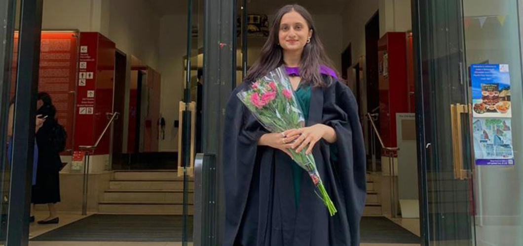 Ariba Fatima stands in front of LSE's Old Building entrance in her graduation robes. She holds a bouquet of flowers in her hands