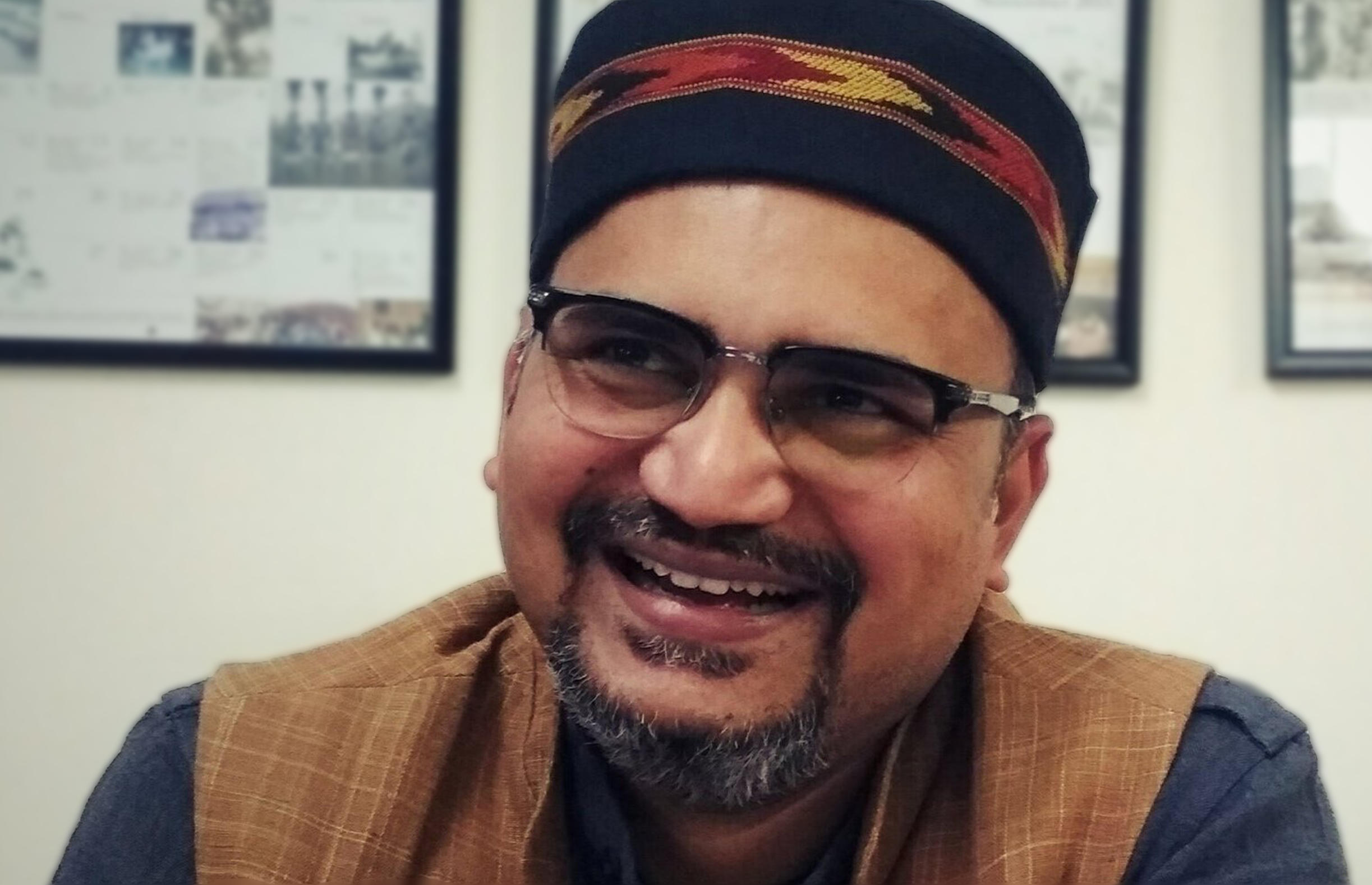 Madhuresh Kumar sit in a room wearing a dark blue and brown top, glases and a Kufi hat. He looks leftwards and smiles.